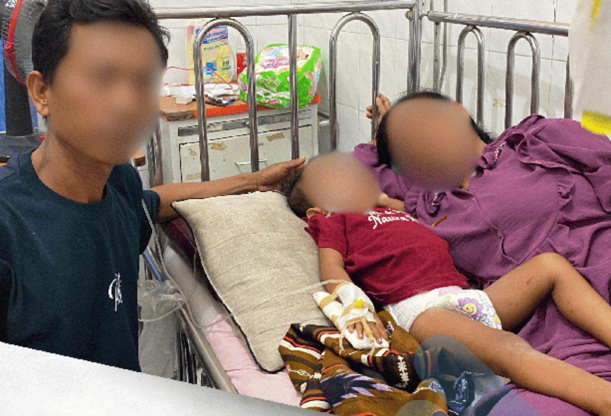 anak umur 2 tahun dengan tangan kanan diinfus berbaring di antara ayah ibunya