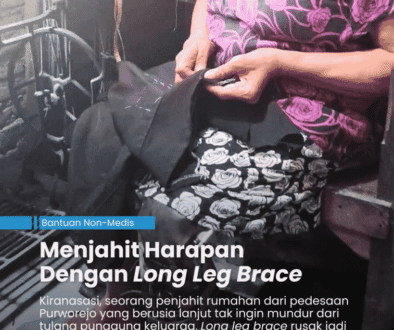 Penjahit perempuan yang sudah tua sedang duduk di kursi yang berada di depan mesih jahit manual sambil memegang celana untuk dijahit