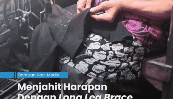 Penjahit perempuan yang sudah tua sedang duduk di kursi yang berada di depan mesih jahit manual sambil memegang celana untuk dijahit