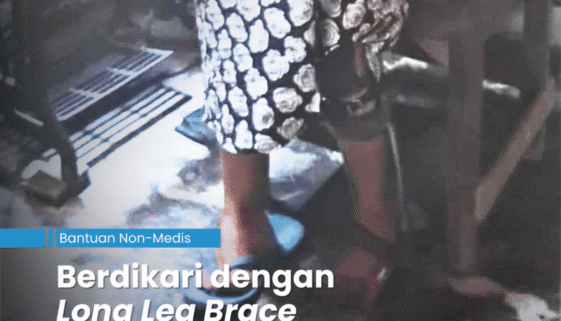 Penjahit perempuan yang sudah tua sedang berdiri di depan mesih jahit manual sambil berpegangan sandaran kursi dengan tangan kiri karena log leg brace yang menopang kakinya sudah rusak dan ingin diganti dengan cara galang dana Bijisesawi.